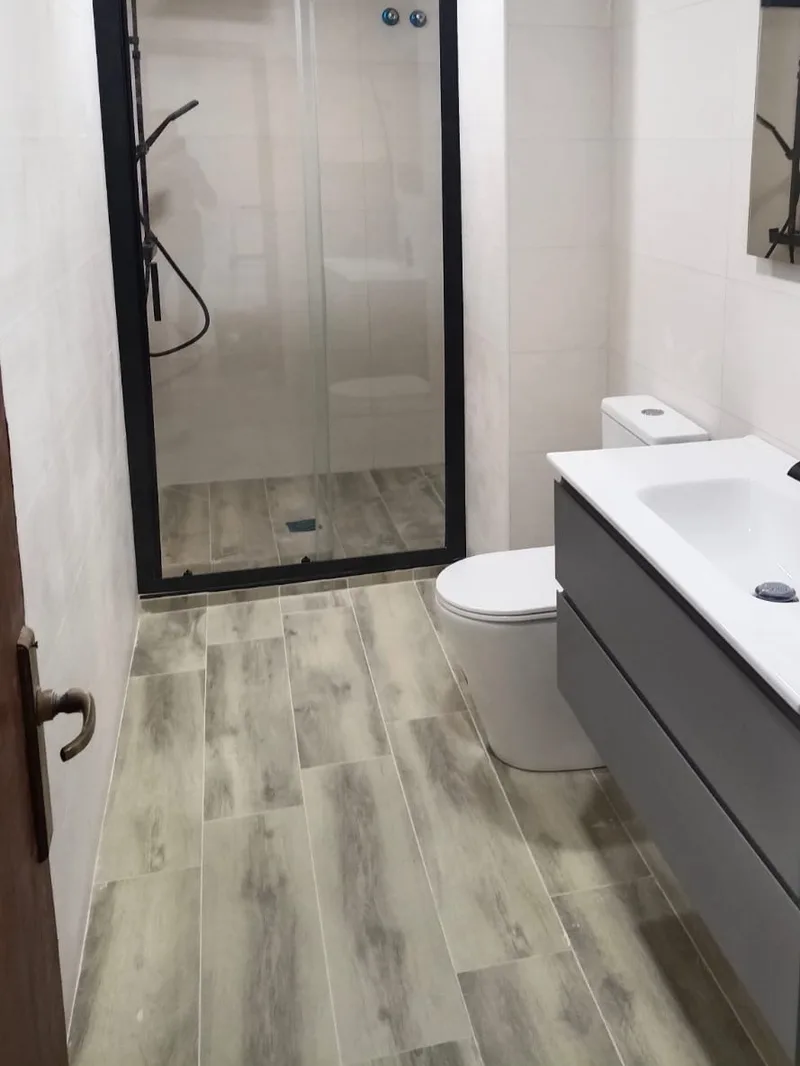 Después: Baño terminado con mampara negra, mueble suspendido con lavabo, inodoro y suelo efecto madera