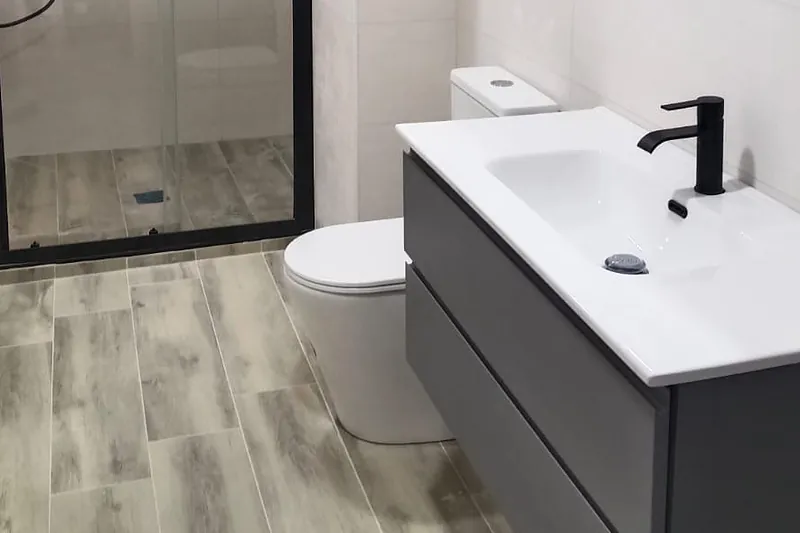 Detalle de baño terminado: lavabo con grifería negra, mueble suspendido gris y suelo porcelánico imitación madera