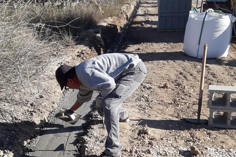 Operario nivelando el hormigón de una zanja de cimentación en una parcela, con bloques al fondo