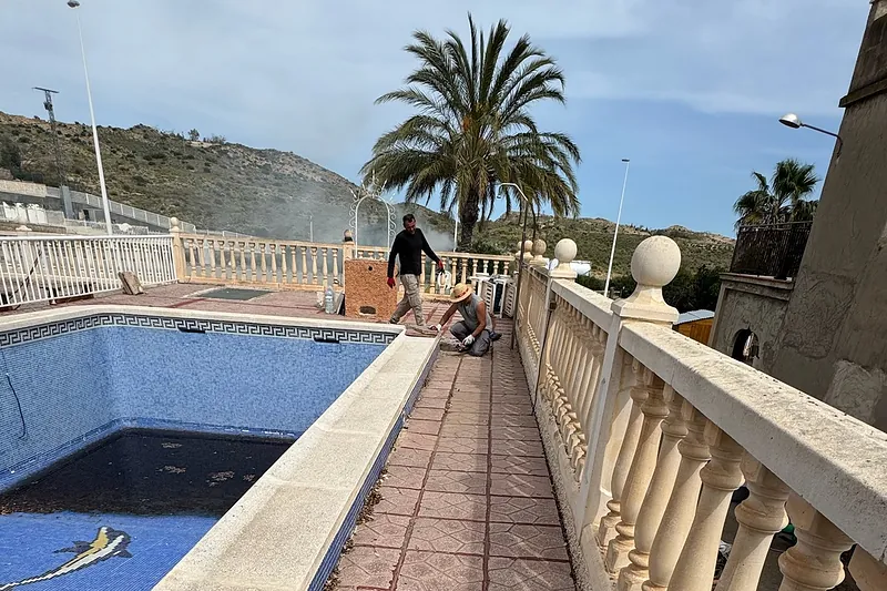 Dos operarios renovando la coronación de una piscina junto a una balaustrada, con una palmera al fondo