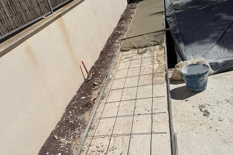 Solera armada con mallazo y mortero recién extendido junto a un muro, con regla y cubo de obra